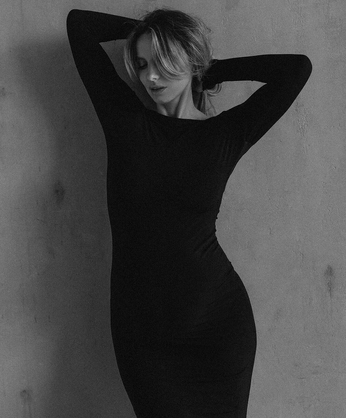 Woman in black dress posing against wall.